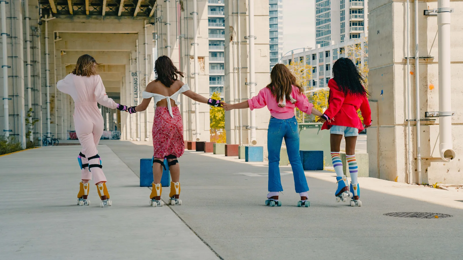 Four women from different backgrounds holding hands while roller skating beneath the Gardiner at The Bentway in Toronto