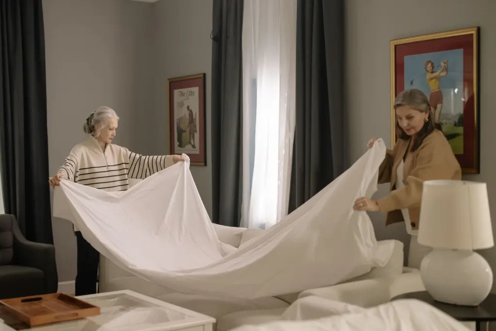 Two women holding a large white sheet while making a bed together