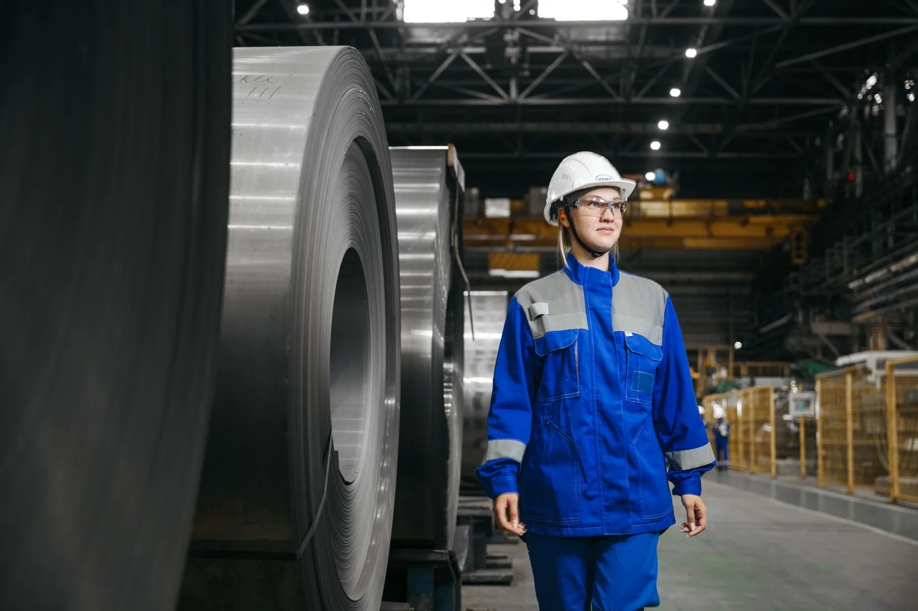 Woman wearing safety gear beside large steel coils in a factory