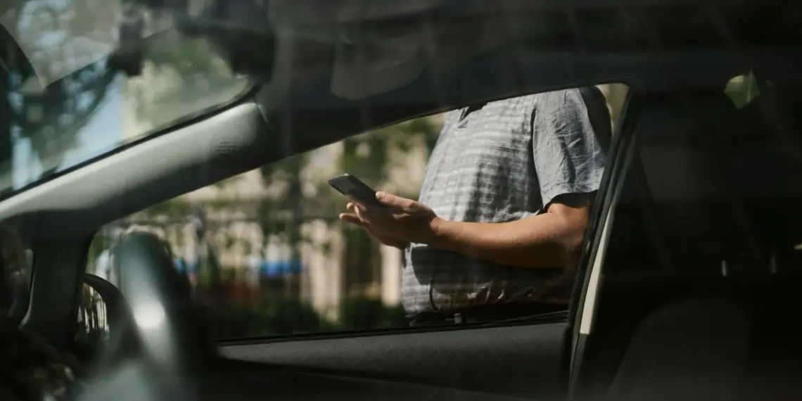 Person checking a smartphone beside a car while waiting for a ride