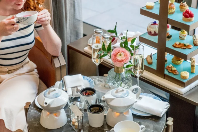 A woman with a teacup beside flowers and a tiered afternoon tea stand at Astor Lounge