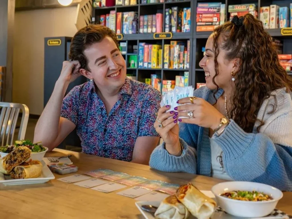 Two women playing a card game with food and drinks at Snakes and Lattes
