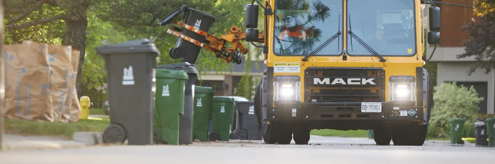 A Toronto collection truck lifting a garbage bin beside green bins and yard-waste bags