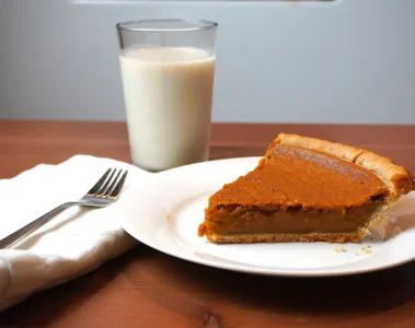 A slice of pumpkin pie on a white plate beside a glass