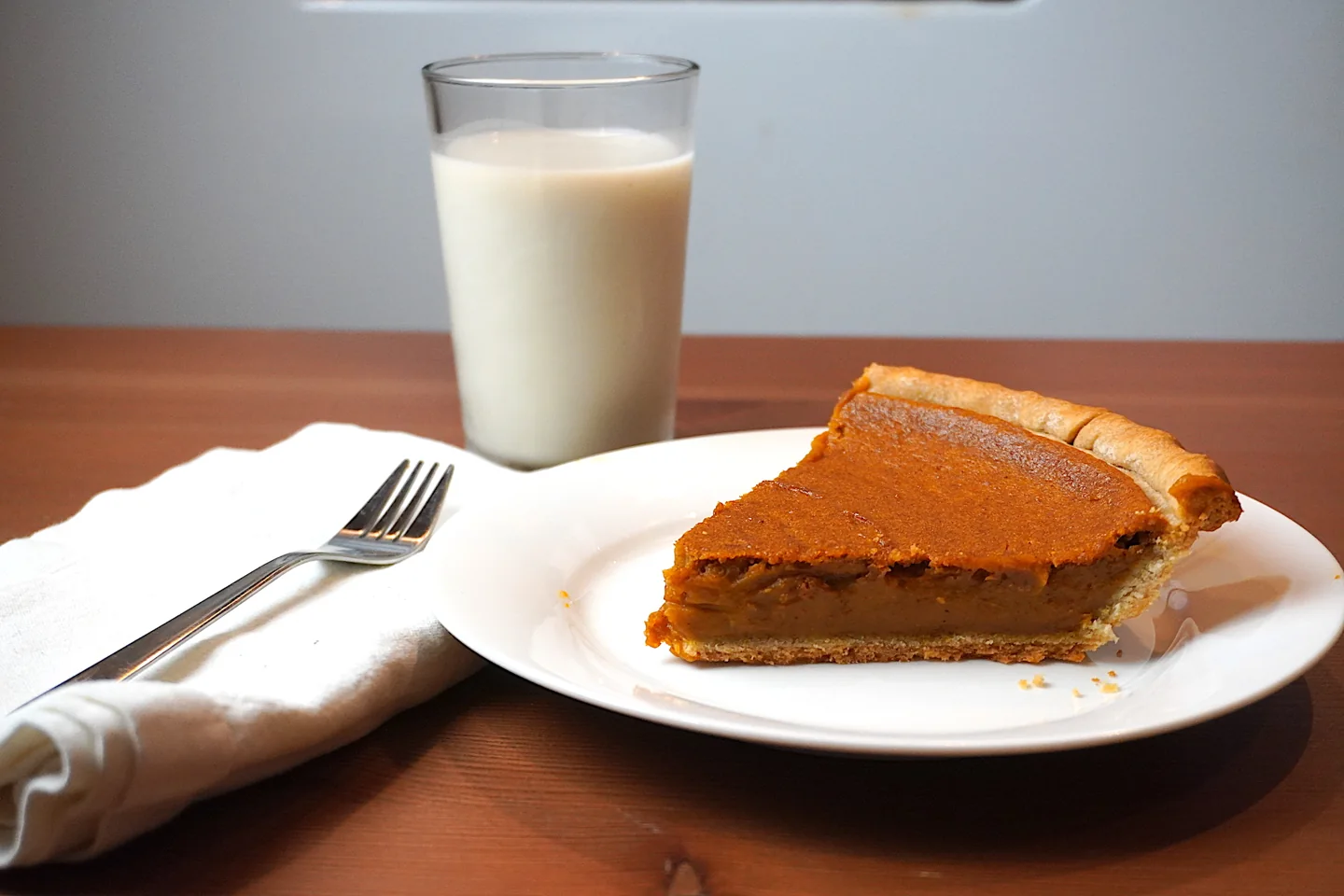 A slice of pumpkin pie on a white plate beside a glass