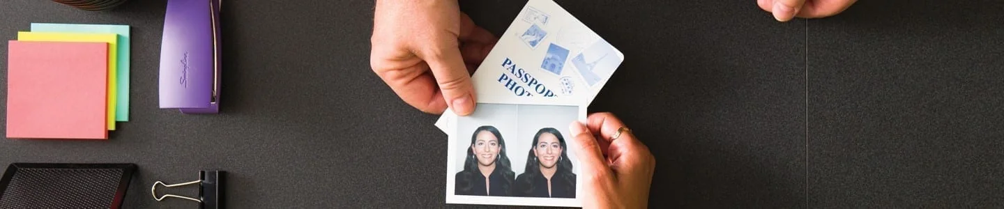 Hands hold a pair of passport photos beside a passport on a desk