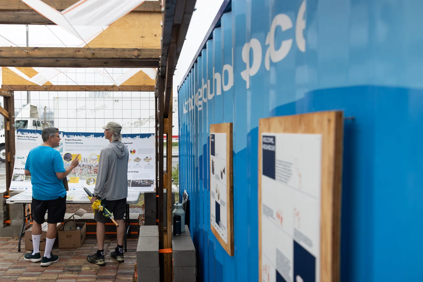 People sit beneath a timber shade pavilion beside a blue shipping container