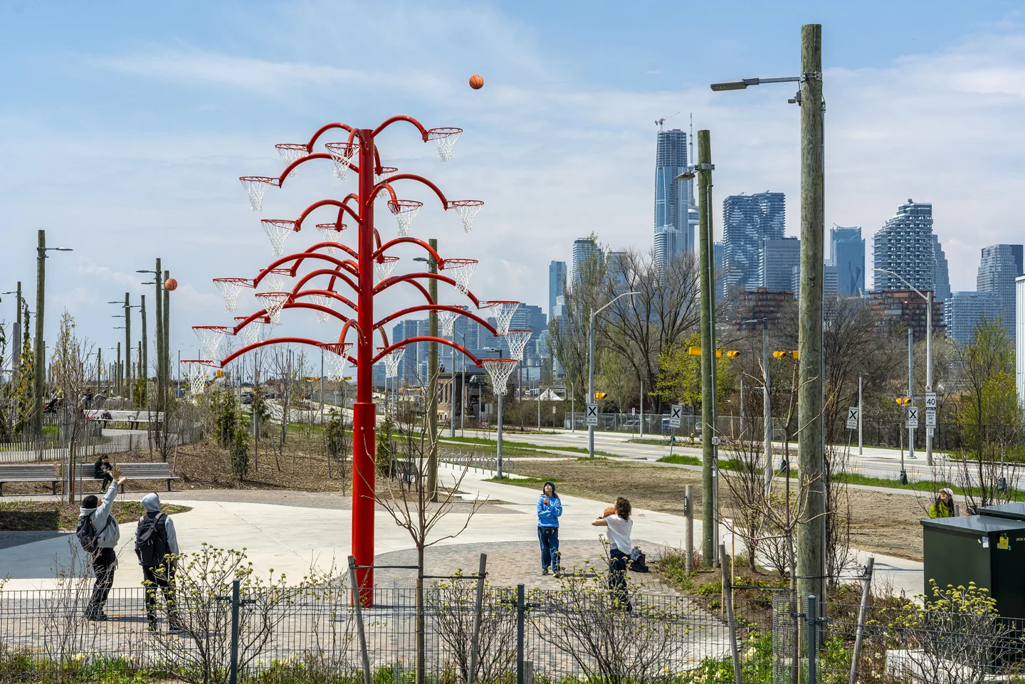 People play basketball beneath a red tree-shaped sculpture with several hoops