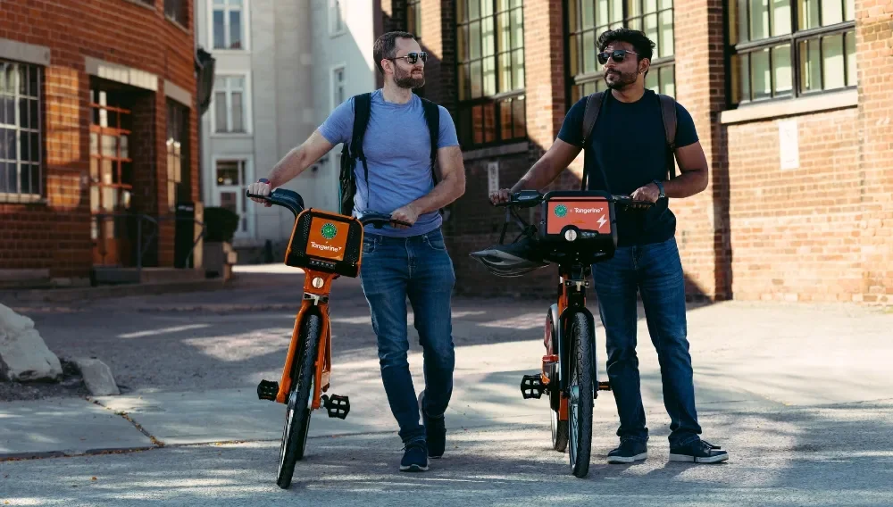 Two riders with backpacks walk orange Bike Share bicycles beside a brick building