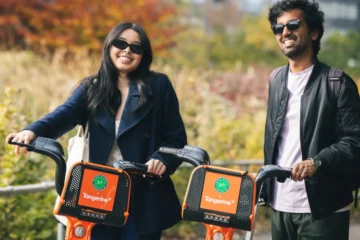 Two people stand with orange Bike Share Toronto bikes beside autumn trees