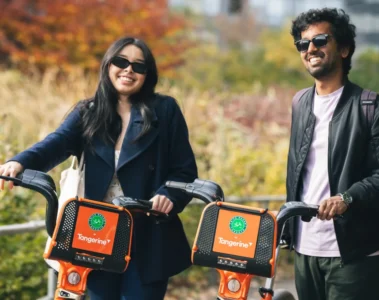 Two people stand with orange Bike Share Toronto bikes beside autumn trees