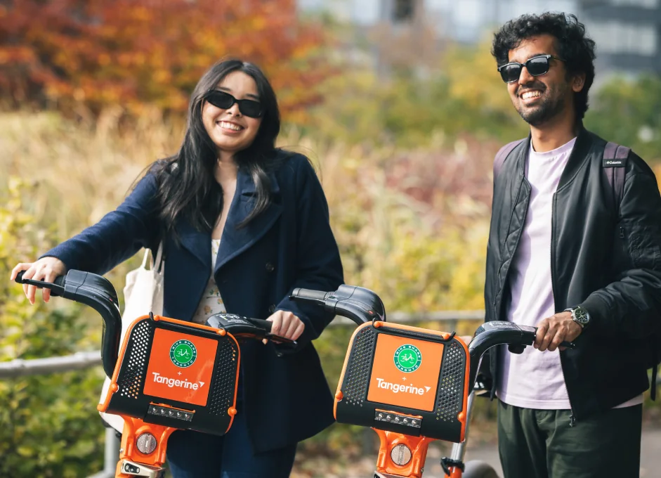 Two people stand with orange Bike Share Toronto bikes beside autumn trees