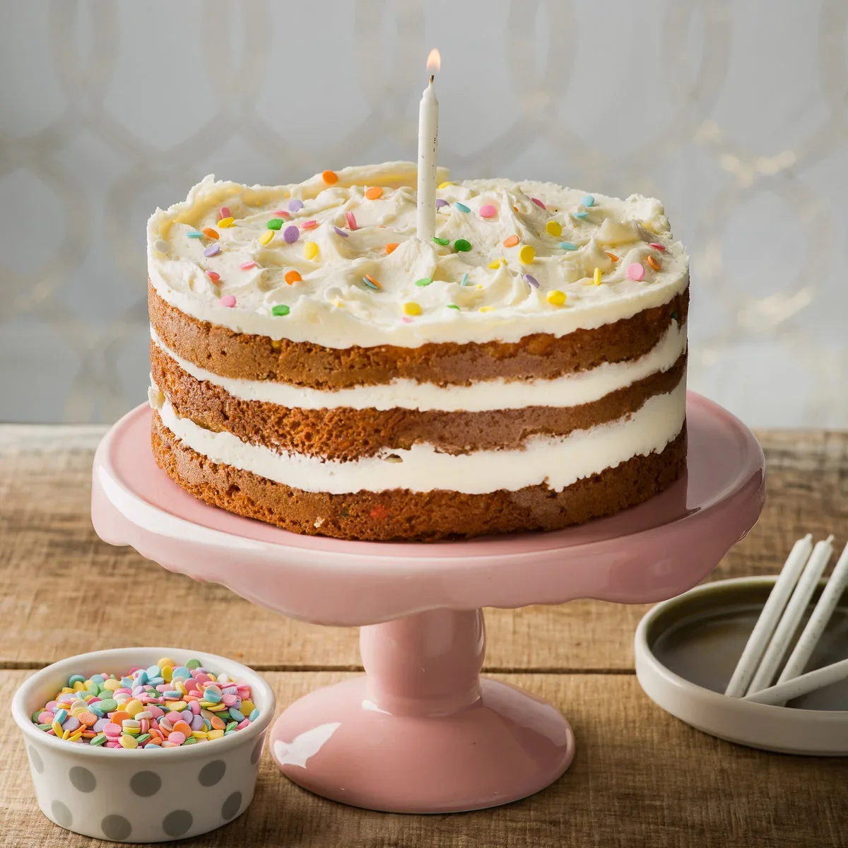 A La Rocca confetti cake on a pink stand with a candle and a bowl of sprinkles