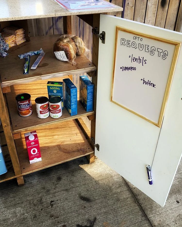 An open community pantry has food on its wooden shelves and a small needs board