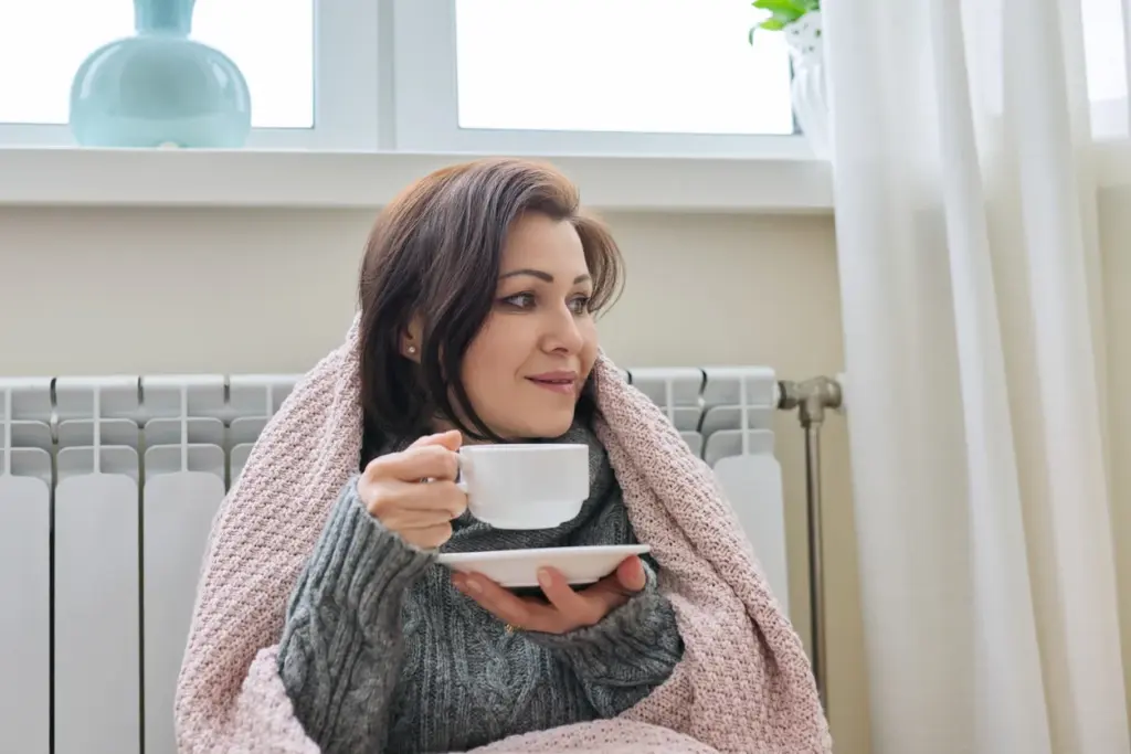 A woman sits beside a radiator with a blanket and hot drink.