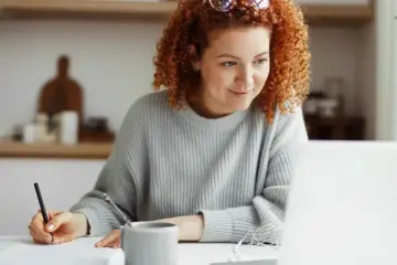 Woman sitting at a table taking notes while looking at her laptop monitor.