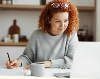 Woman sitting at a table taking notes while looking at her laptop monitor.