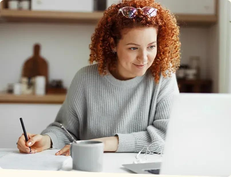 Woman sitting at a table taking notes while looking at her laptop monitor.
