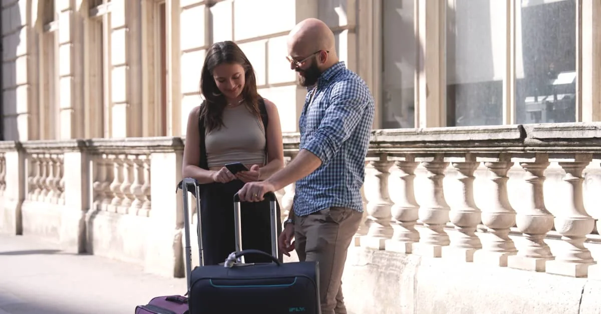 Two travellers stand with luggage while looking at a phone