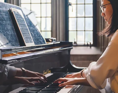 A woman plays a grand piano beside a sunlit window at Hart House