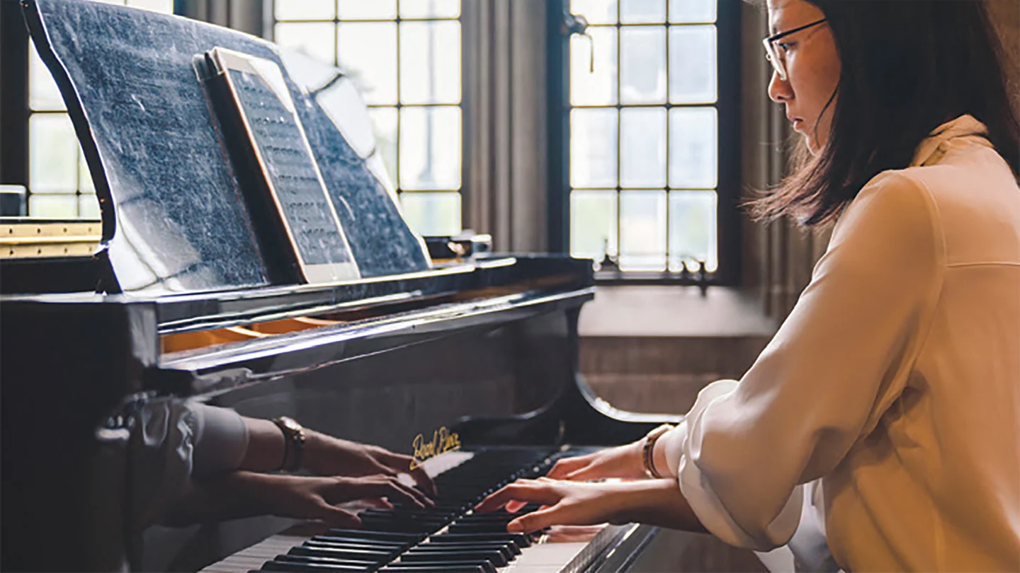 A woman plays a grand piano beside a sunlit window at Hart House
