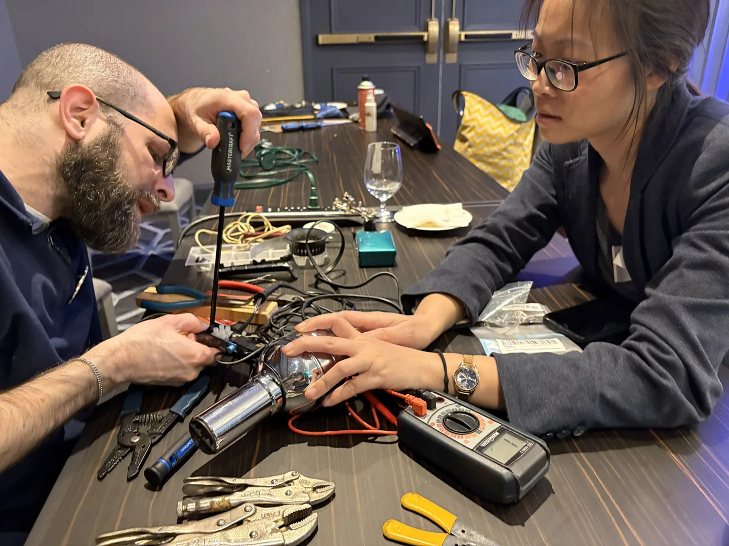 A fixer and a visitor examine electronics and a small appliance at a table
