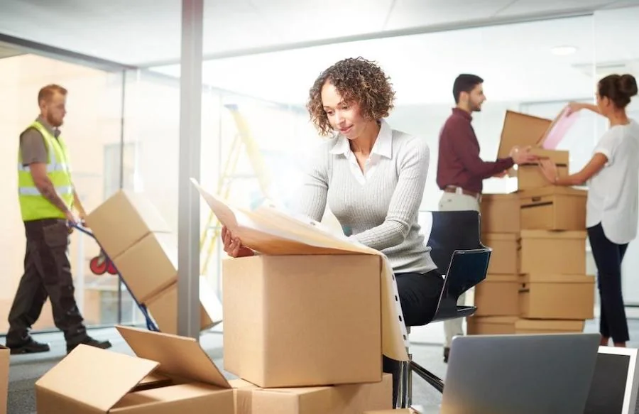 A woman closes a packed box while movers carry household items in the background