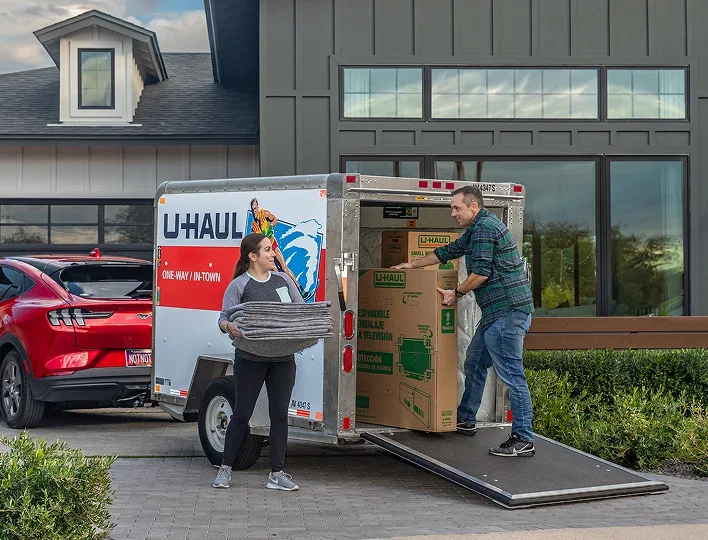 A woman carries folded moving blankets while a man loads boxes into a U-Haul trailer