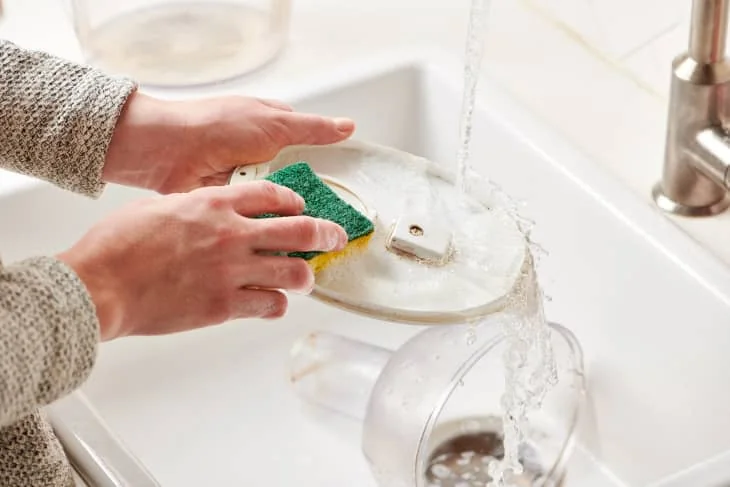 A person washes a pitcher lid with a sponge beside a kitchen sink