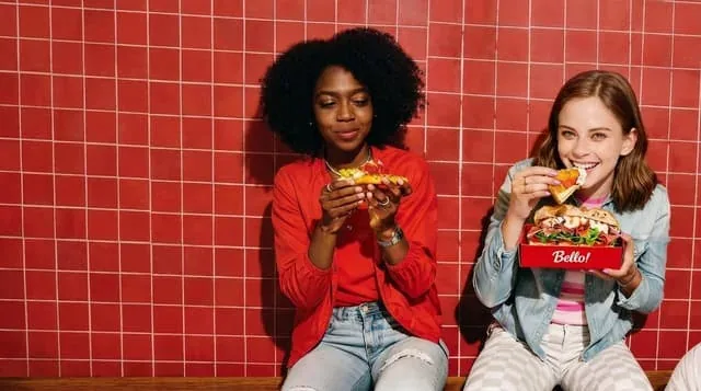 Two women eating sandwiches in front of a red tiled wall
