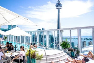 Guests relax beside Bisha Hotel Toronto's rooftop pool with the CN Tower, Rogers Centre and Lake Ontario beyond