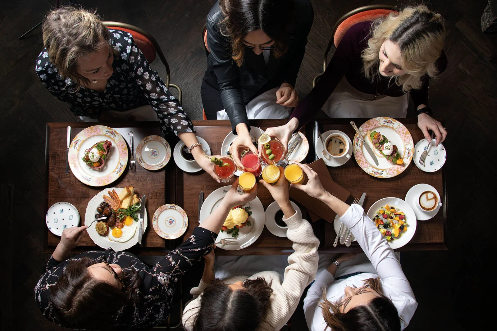 An overhead shot of six friends cheersing mimosas and Caesars over a table of brunch food at Maison Selby