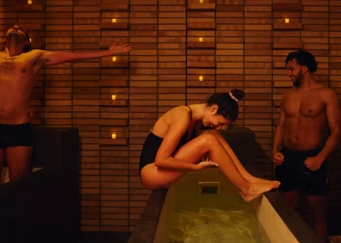 Three people in swimwear enjoy a spa experience, with one woman sitting on the edge of an ice bath, smiling, while the others react playfully