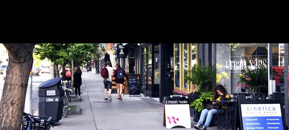 People walk beside shops and trees on Danforth Avenue