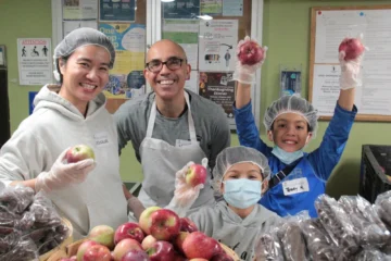 Smiling adults and children holding apples while volunteering at Good Shepherd’s Thanksgiving meal